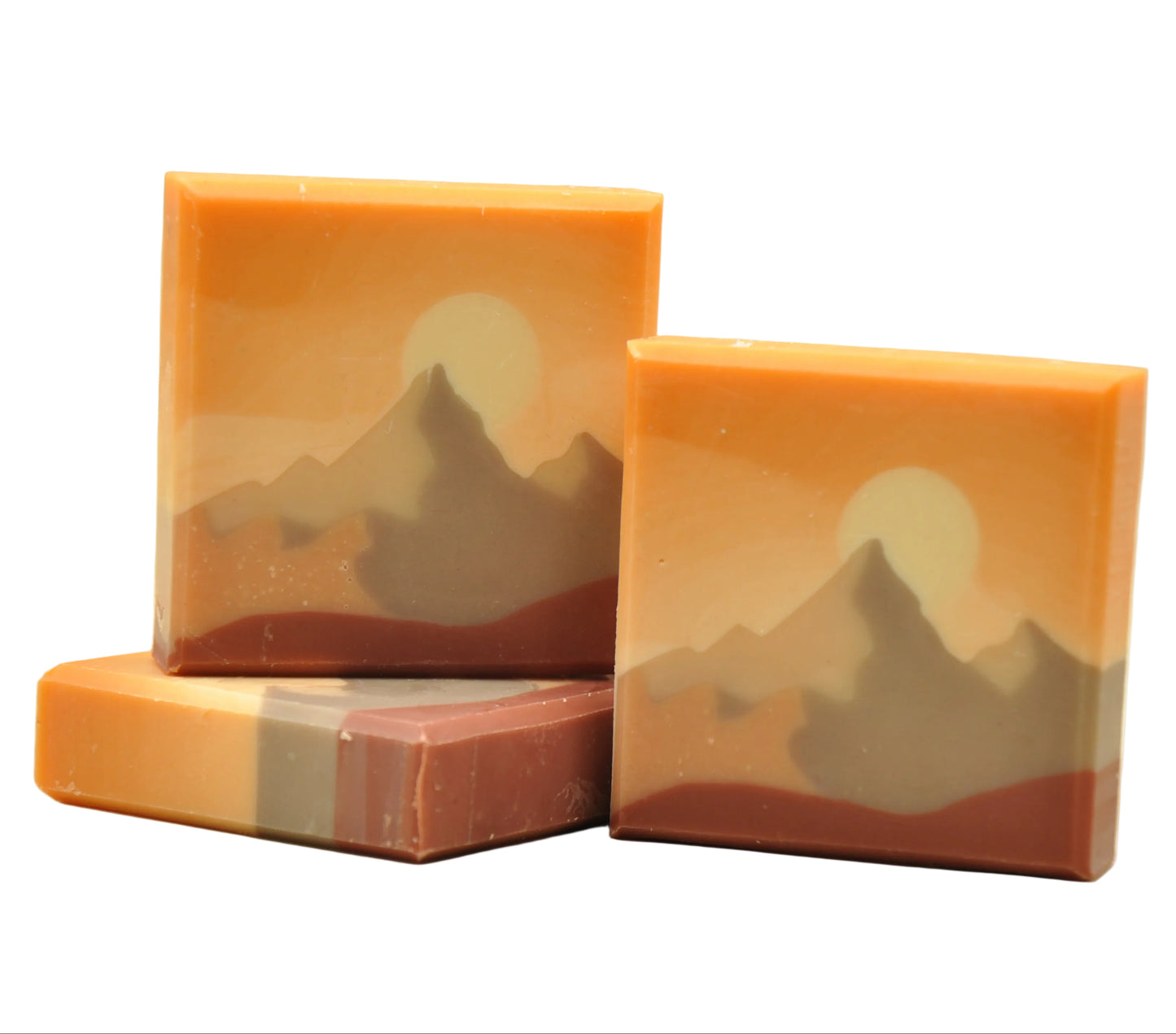 Drei Stücke handgemachte Kaltverseifungsseife namens Everest, mit einem Berglandschafts-Design in warmen Amber- und Terrakotta-Tönen, einer cremefarbenen Sonne hinter grau-taupefarbenen Berggipfeln und einer tiefen Terrakotta-Basisschicht, duftend nach Zitrus und Kiefer.