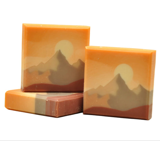 Drei Stücke handgemachte Kaltverseifungsseife namens Everest, mit einem Berglandschafts-Design in warmen Amber- und Terrakotta-Tönen, einer cremefarbenen Sonne hinter grau-taupefarbenen Berggipfeln und einer tiefen Terrakotta-Basisschicht, duftend nach Zitrus und Kiefer.