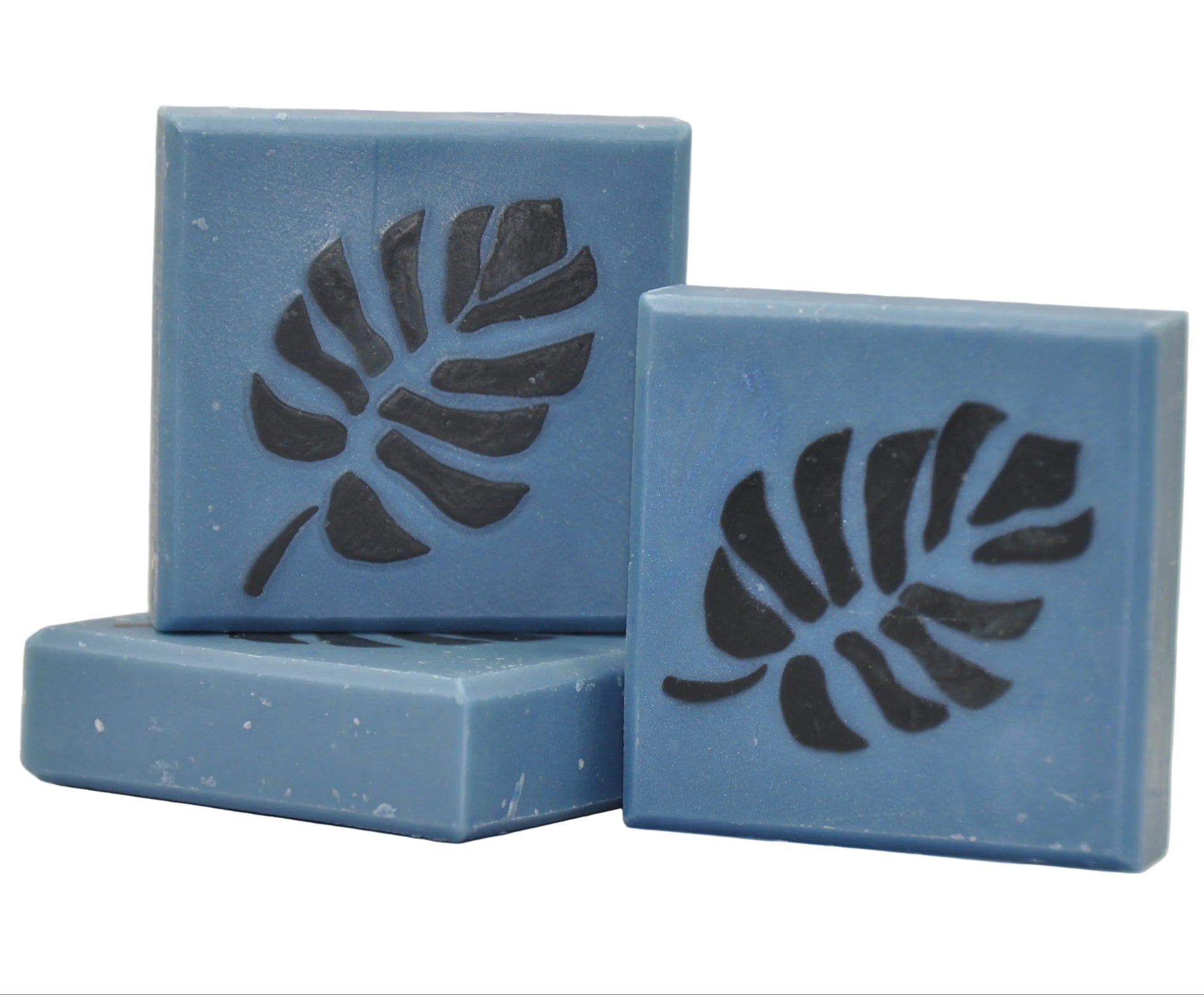 „Monstera Soap on Blue“ – drei stahlblaue, handgemachte Seifenstücke mit einem dunkelgrauen Monstera-Blattmuster, das sich durch die Seife zieht, präsentiert auf weißem Hintergrund.
